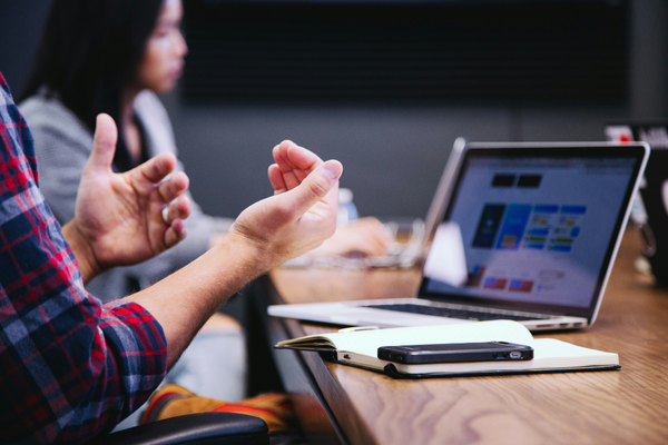 Close up of person meeting with others around a conference table, gesturing with hands with a laptop nearby.