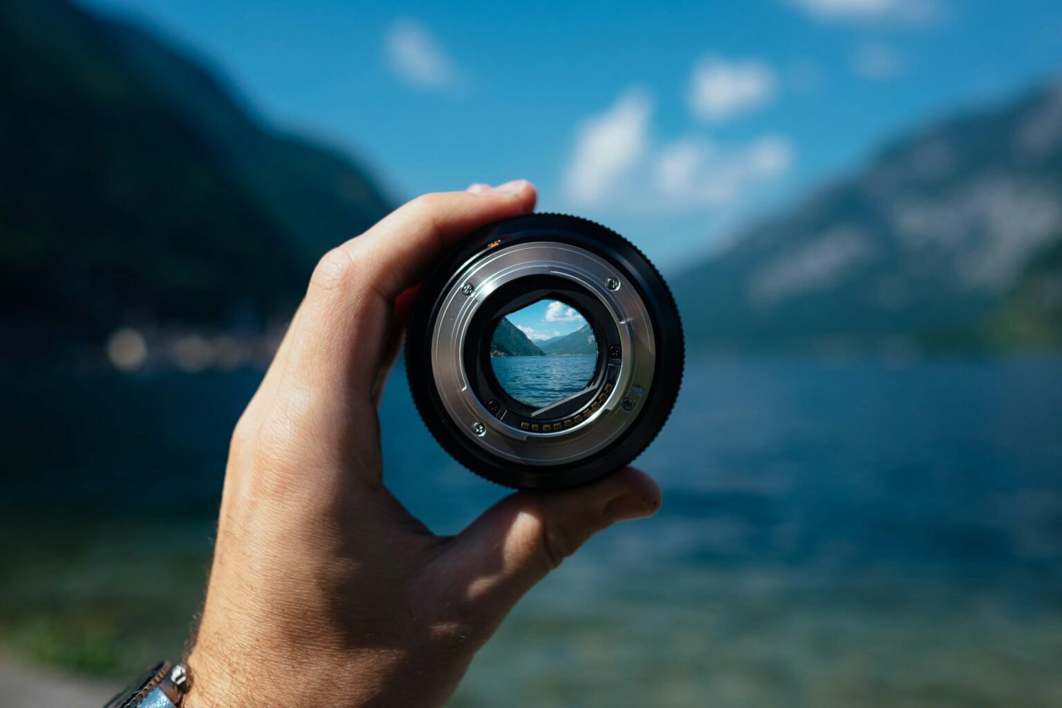 A hand holding a camera lens in front of a scenic lake and mountains in the background