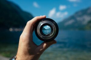 A hand holding a camera lens in front of a scenic lake and mountains in the background