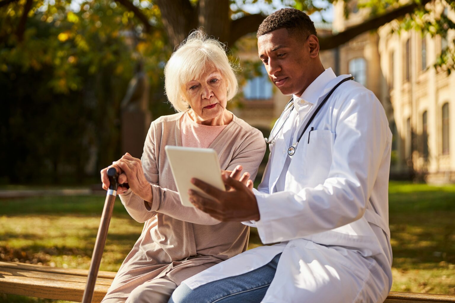 A older woman sitting on a bench holding onto a cane consulting with her doctor, while looking at a digital tablet together.