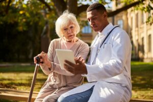 A older woman sitting on a bench holding onto a cane consulting with her doctor, while looking at a digital tablet together.
