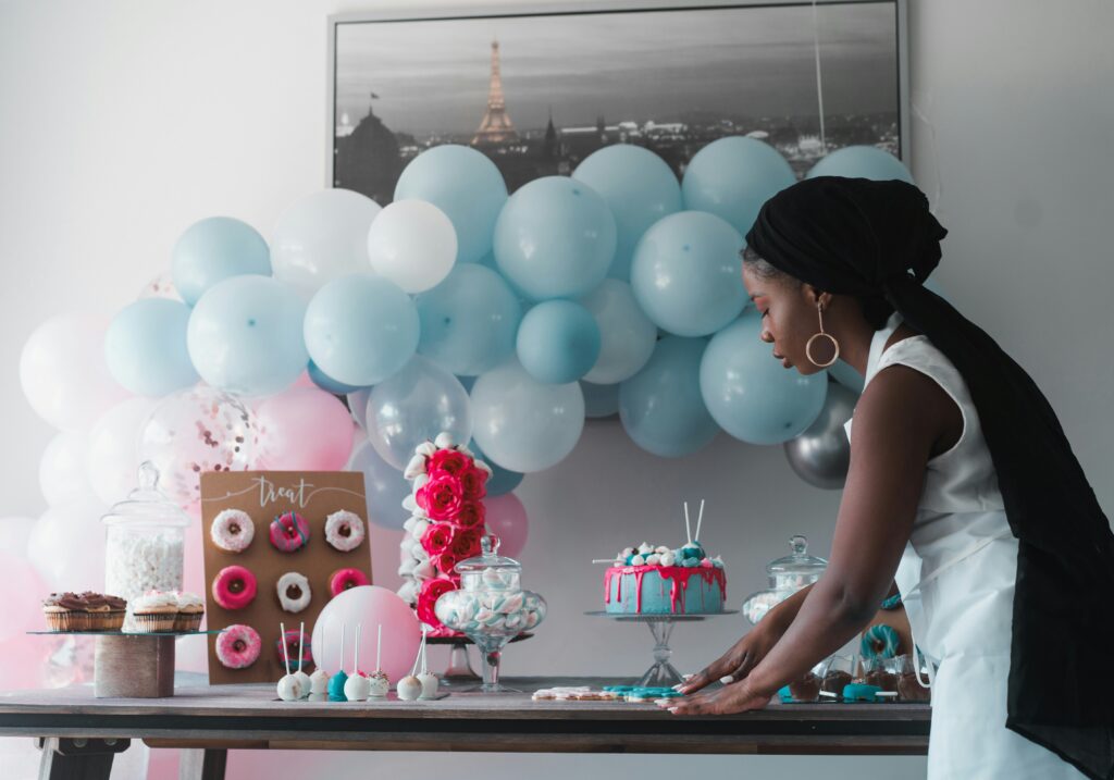 Black woman in white dress decorating a table for a celebratory party with blue balloons and a cake, among other decorations.