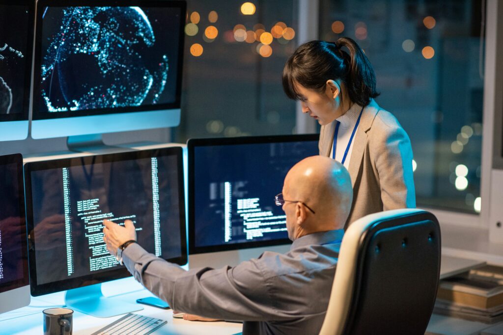 A male scientist is sitting at a desk pointing to data on his screen to a female colleague, who is standing to his right.