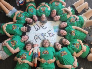 Campers from Oscar's Teen MS Camp lying in a circle around a sign that reads "We are," wearing matching camp shirts