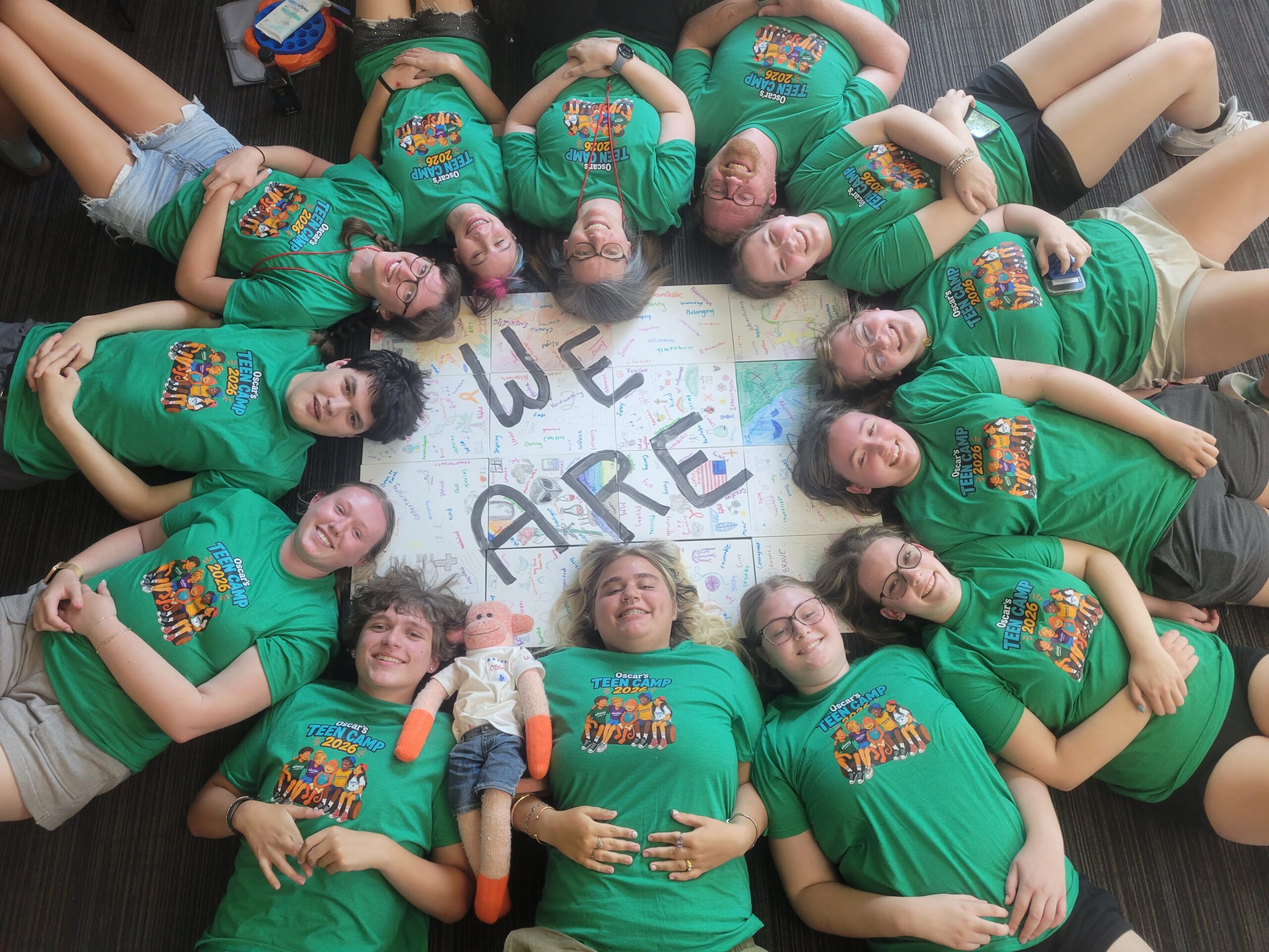 Campers from Oscar's Teen MS Camp lying in a circle around a sign that reads "We are," wearing matching camp shirts
