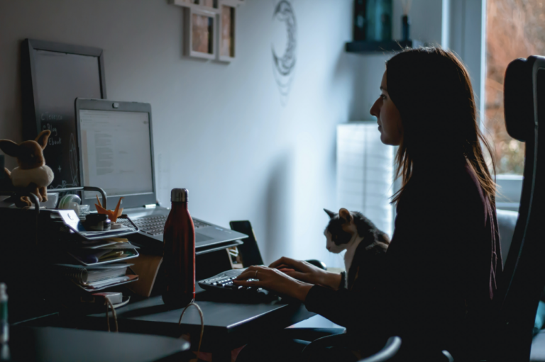 Woman sitting at her desk at home working on computer with a cat on her lap.